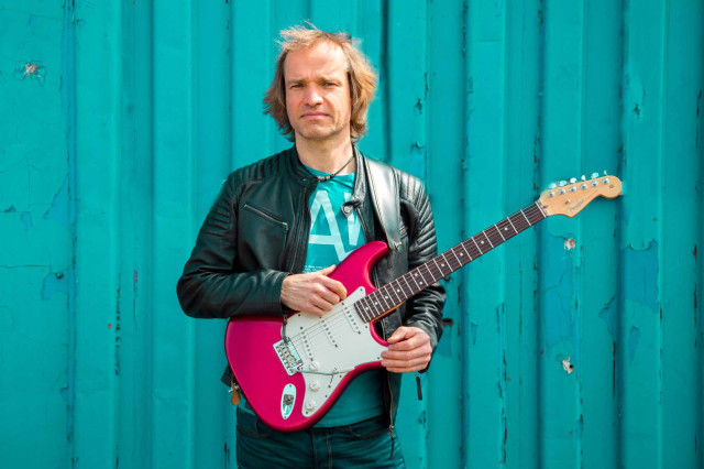 Thorsten Praest - Der Gitarrist Thorsten Praest steht vor einer blauen Wand im Tageslicht und hält seine Gitarre in der Hand