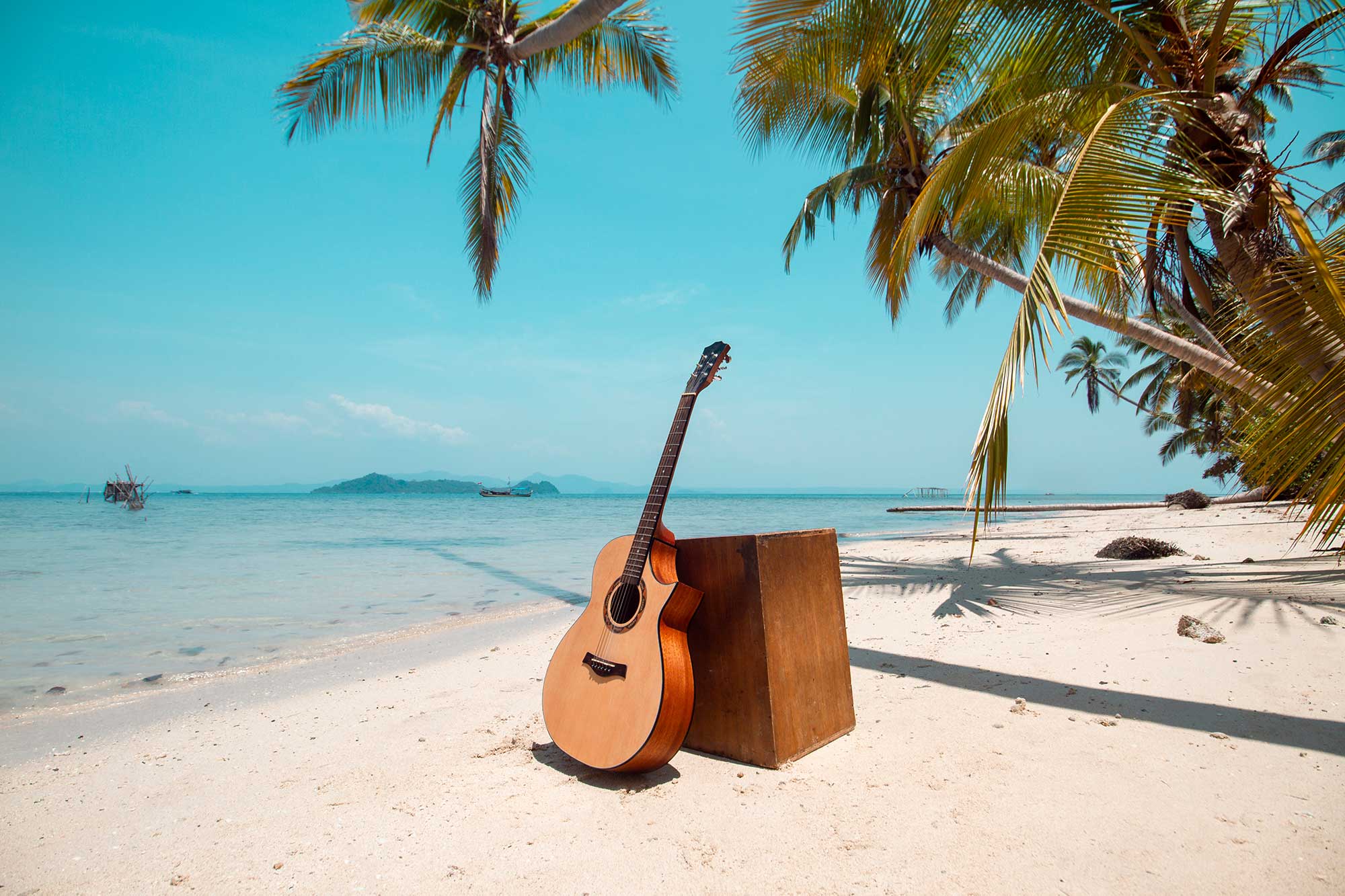 Eine Cajon mit angelehnter Gitarre auf dem Sandstrand mit Blick auf eine Bucht mit blau schimmerndem Meer