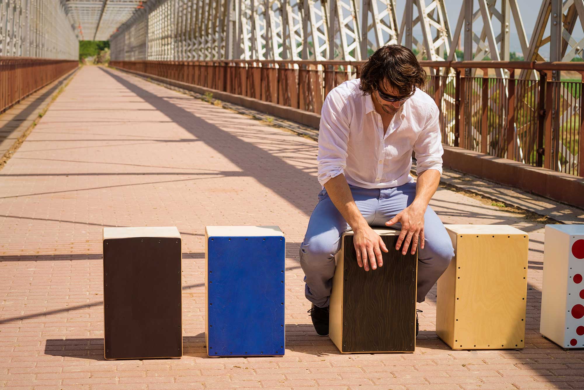 Ein Mann mit Sonnenbrille spielt auf einer Bruecke bei strahlendem Sonnenschein auf einer Cajon